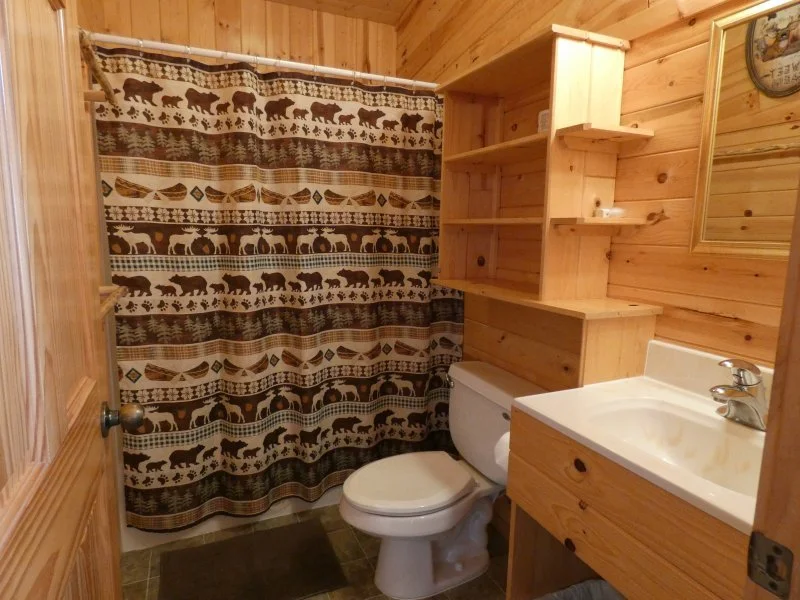Small rustic bathroom with wooden walls and ceiling, a patterned shower curtain featuring bears and trees, a toilet, a wooden vanity with a sink, a wall clock, and open wooden shelves.