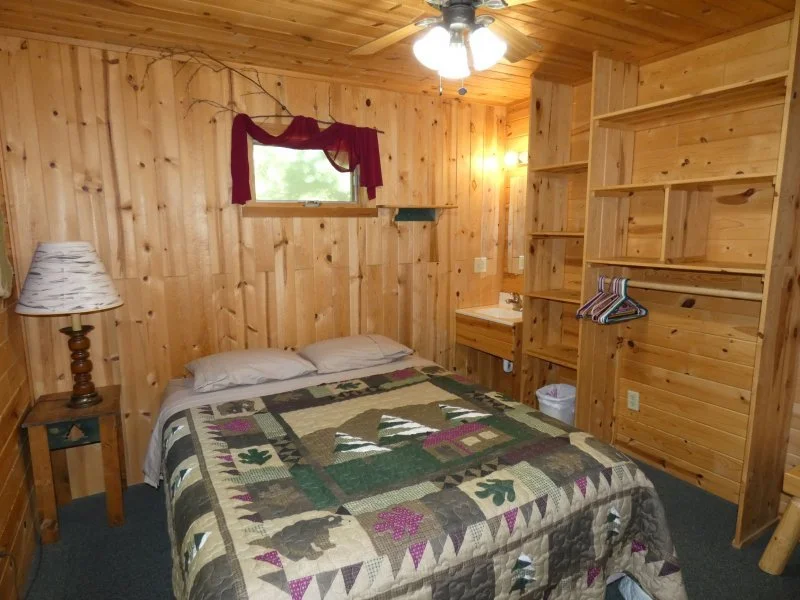 A cozy bedroom with wood-paneled walls, a bed with a colorful quilt, a nightstand with a lamp, a small window with a red curtain, and a built-in open wooden closet.