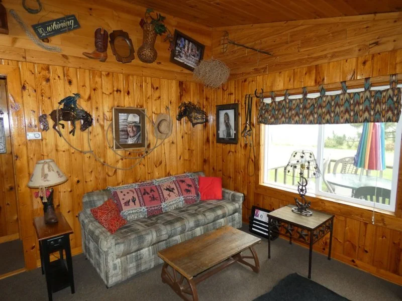 A cozy cabin living room with wood-paneled walls, a plaid sofa with decorative pillows, a side table with a lamp, a small table with a lamp near a large window with colorful outdoor cushions, and various decorative items on the walls including pictures, signs, and hanging ornaments.