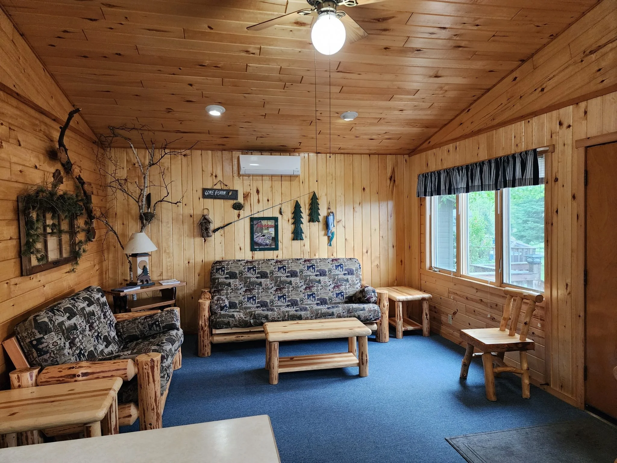 Interior of a log cabin living room with wooden walls and ceiling, featuring a ceiling fan with a light, a window with black and white striped curtains, log furniture including two sofas with patterned cushions, a coffee table, a side table with a lamp, and various wall decorations including a sign, framed picture, and hanging evergreen decorations.