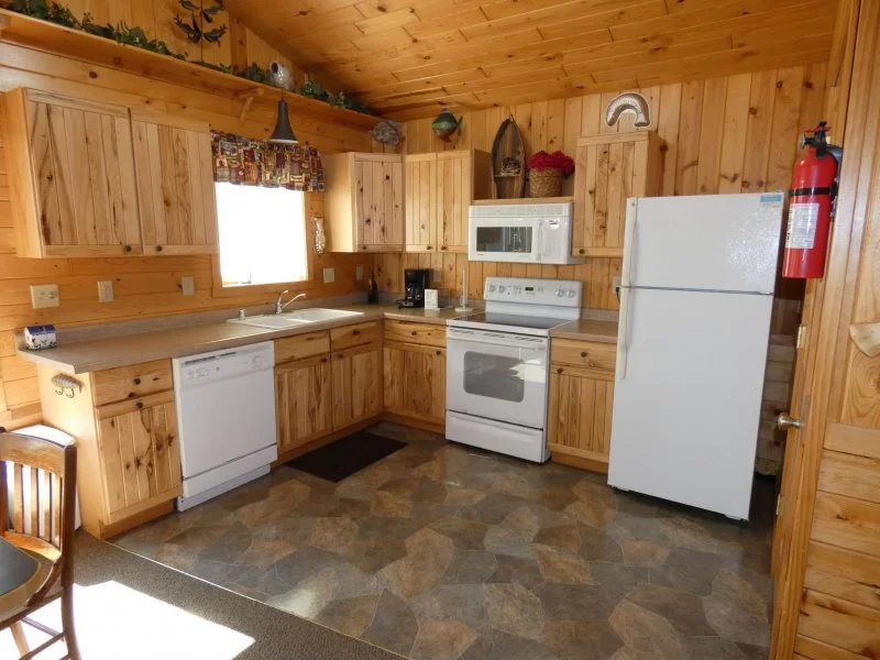 Wood-paneled kitchen with white appliances, including a dishwasher, stove, microwave, and refrigerator, with a window above the sink, and a fire extinguisher on the wall.