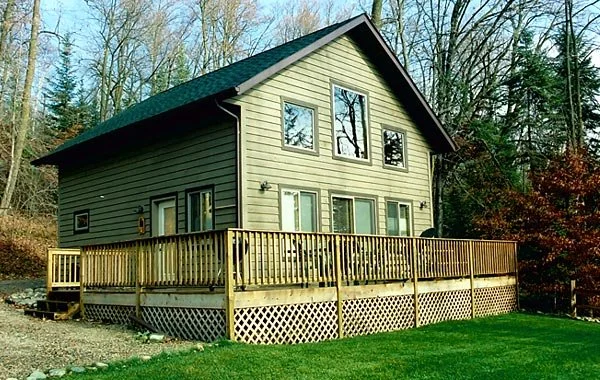 A two-story house with beige siding and a dark roof, surrounded by trees. It has multiple windows and a wooden deck in the backyard with a lattice skirt.
