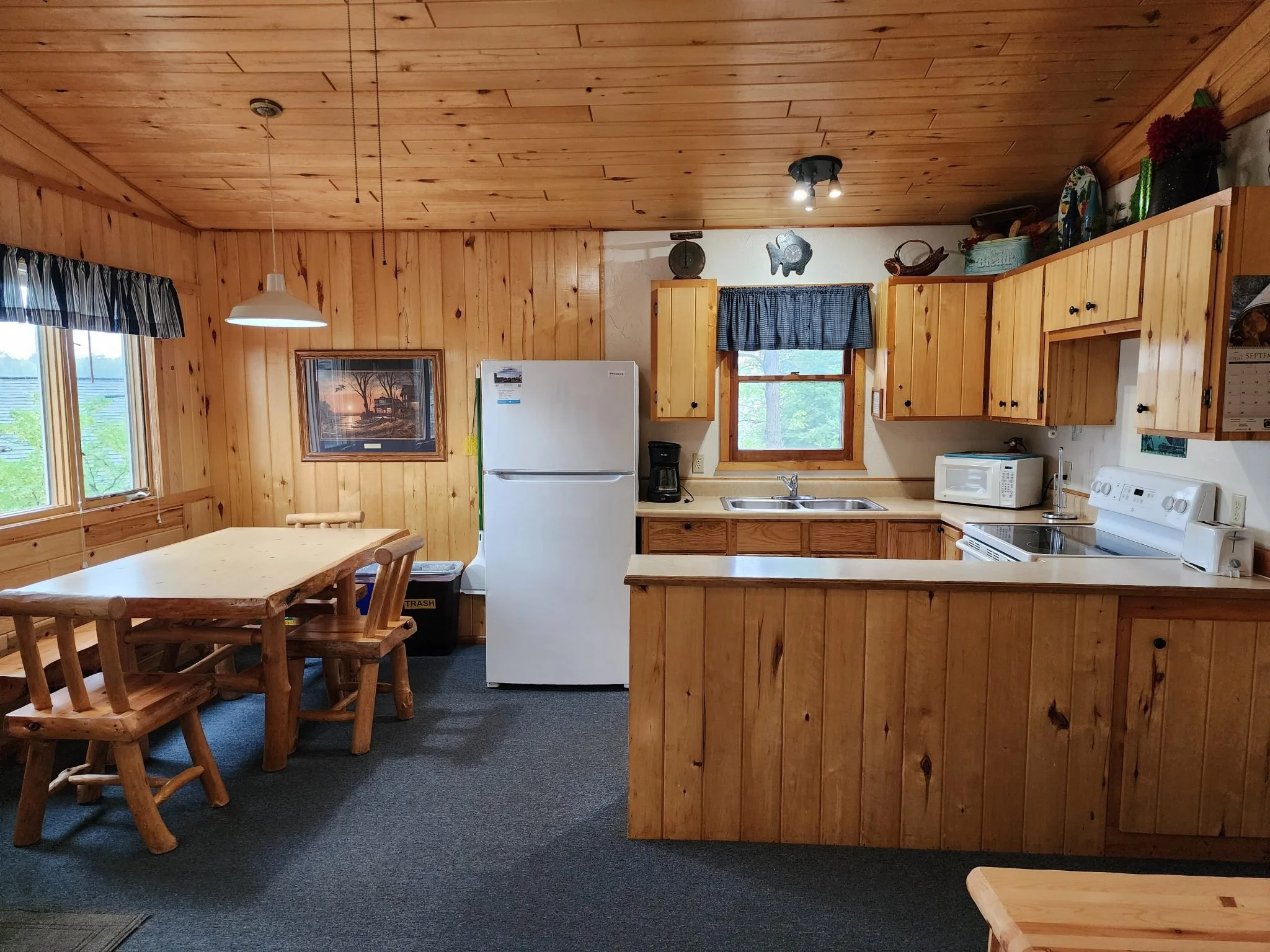 A wooden kitchen with natural wood cabinets, ceiling, and walls, a white refrigerator, microwave, and stove, with windows and wall decorations.