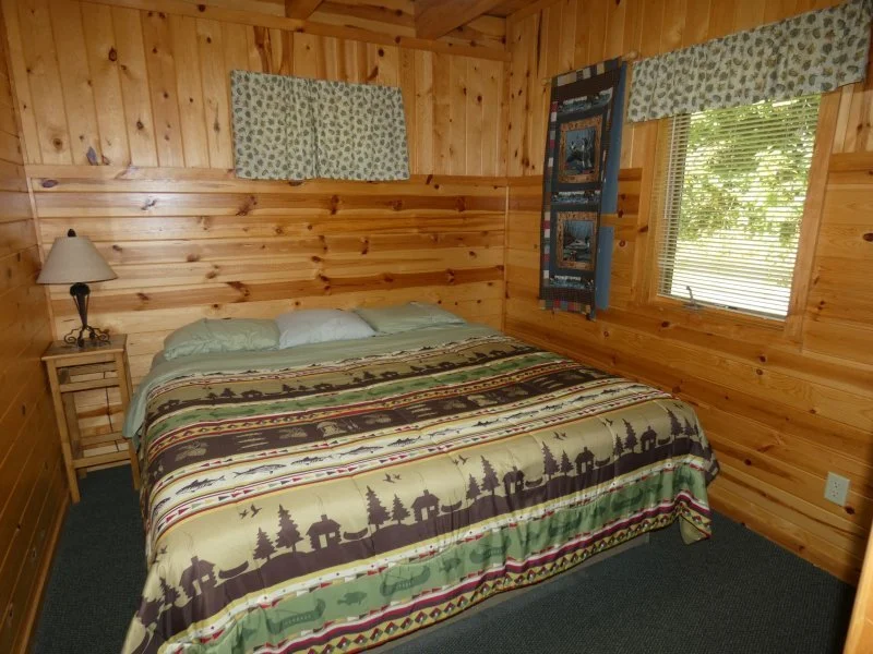 A cozy bedroom with wooden walls, a large bed with patterned bedspread, a nightstand with a lamp, a window with blinds, and a fabric wall hanging.