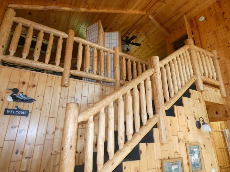 Wood interior with a staircase made of log-style wood railing, leading up to a loft with a wooden ceiling and walls, ceiling fan, wall-mounted light, and framed pictures.