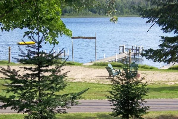 A lakeside scene with a sandy beach, green grass, and trees, featuring a wooden dock, a yellow kayak, and two chairs near the water in the background.