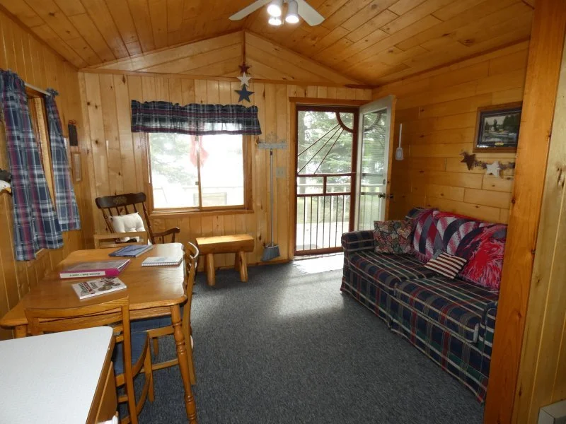 Cozy living room with wood-paneled walls and ceiling, featuring a plaid sofa, a wooden dining table with chairs, a window with plaid curtains, and a small balcony door with a view of trees outside.