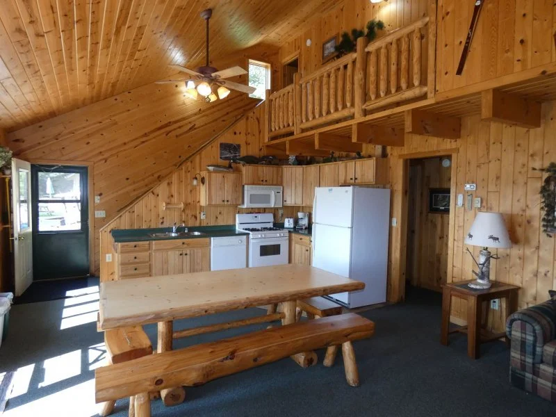 Interior of a wooden cabin kitchen with a wooden dining table and bench, white refrigerator, stove, microwave, and a small side table with a lamp; wood-paneled walls and ceiling, loft area with railing, and sunlight coming through the door window.