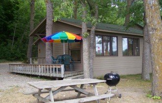 A small house or cabin with large windows, surrounded by trees. In front, there is a wooden picnic table and a patio with a multicolored umbrella, outdoor chairs, and a charcoal grill.