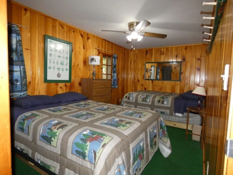 A bedroom with wood-paneled walls, two beds with woodland-themed quilts, a ceiling fan, and various wall and table decorations.