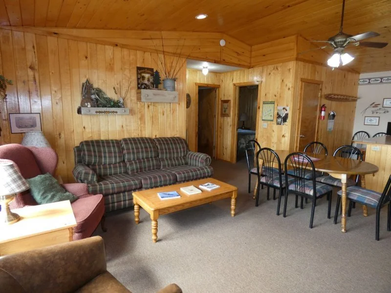Living room with wood-paneled walls, a plaid sofa, a pink armchair, and a wooden coffee table. Adjacent to the living area is a dining table with six chairs. There are ceiling fans and various wall decorations.