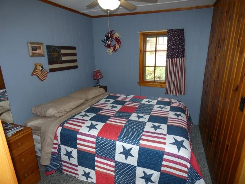 A cozy bedroom with light blue walls, wood paneling on one wall, and a window with patriotic-themed curtains. The bed has a red, white, and blue quilt with stars and stripes, and beige pillows. There are decorative items on the wall, including an American flag, a star flag, and a wreath.