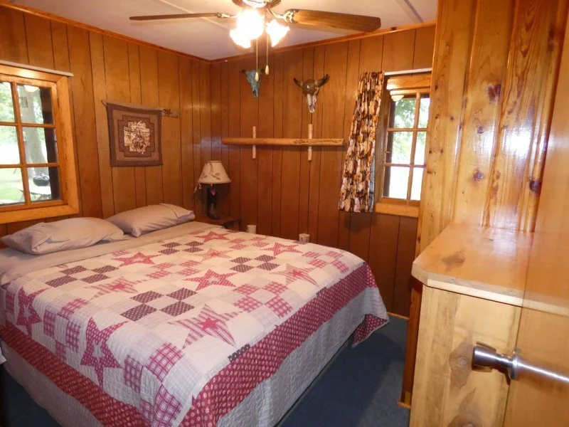 A cozy bedroom with wooden panel walls, two windows with floral curtains, a bed with a quilt featuring red and white patterns, a lampshade on a nightstand, and mounted animal head decorations on the walls.