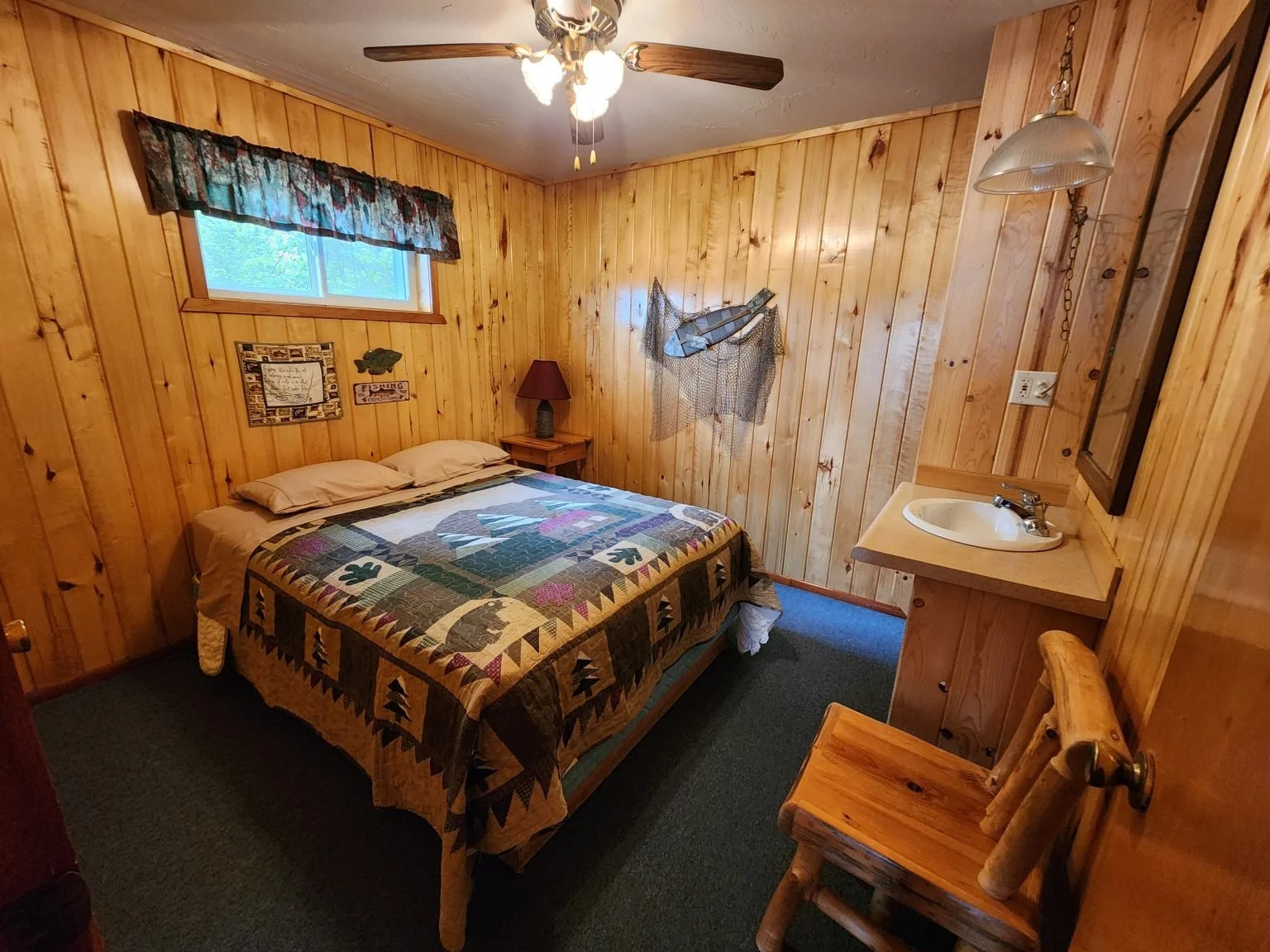 A cozy bedroom with wood-paneled walls, a bed with a patchwork quilt, a small window with floral curtains, a ceiling fan, a rustic wooden chair, a small sink with a mirror, and decorative fishing fishing items on the wall.