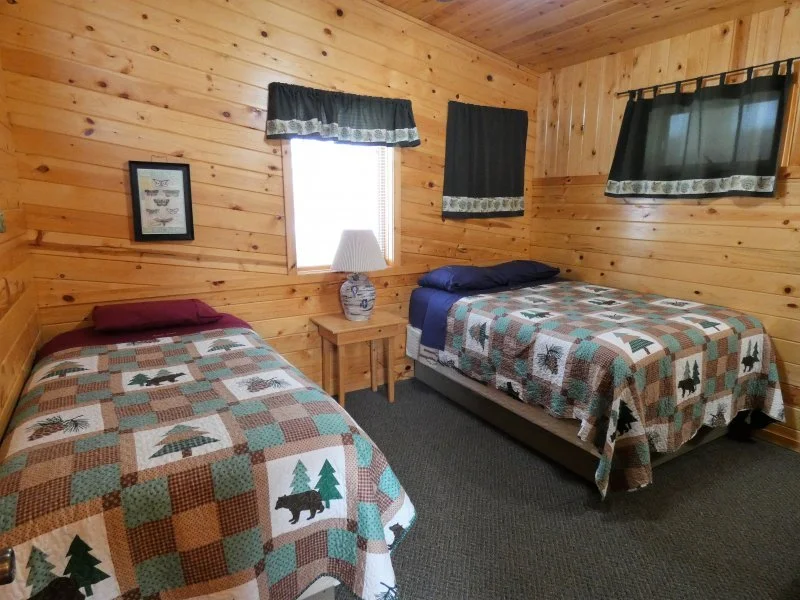 A cozy bedroom with two twin beds covered in quilted bedspreads featuring pine trees and bears, a wooden nightstand with a lamp, wood-paneled walls, and small windows with curtains.