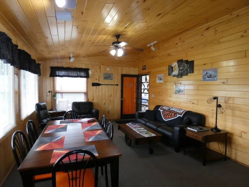 A cozy living and dining room with wood-paneled walls and ceiling, a large wooden table with eight black chairs, a black leather sofa with a Harley-Davidson blanket, a small coffee table with magazines, a side table with a lamp, and various motorcycle-themed wall art.