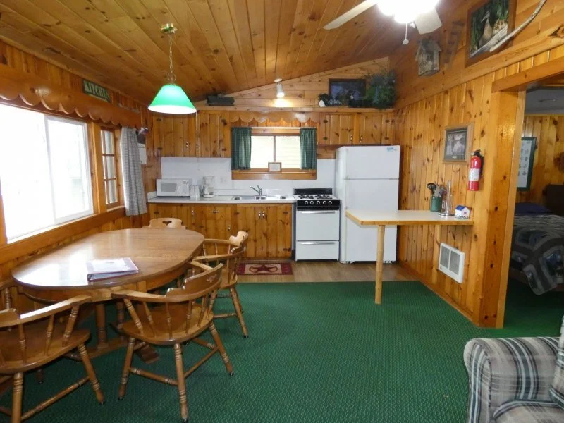 Wood-paneled kitchen and dining area with an oval dining table, four chairs, a refrigerator, oven, microwave, and a window with curtains. A small counter with items and a fire extinguisher with a doorway leading to a bedroom.