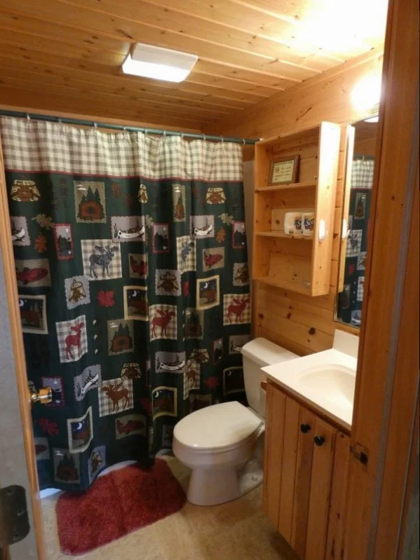 A small bathroom with wood-paneled walls and ceiling. There is a shower with a curtain featuring a rustic, mountain scene. A red rug is on the floor. The bathroom has a white toilet and a wooden vanity with a white sink. Above the toilet is a wooden shelf with picture frames and decorative items.