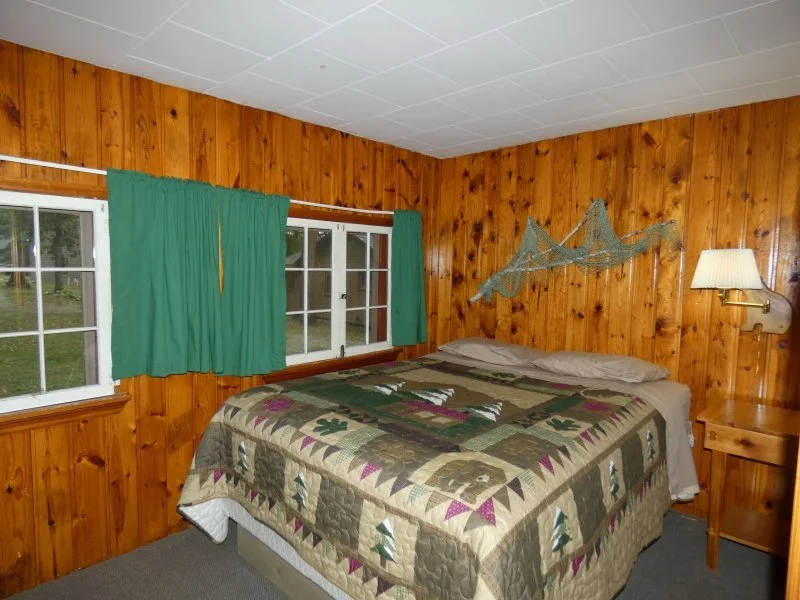 Bedroom with wood-paneled walls, a quilted bedspread, two small windows with green curtains, a wall-mounted lamp, and a decorative fishing net on the wall.