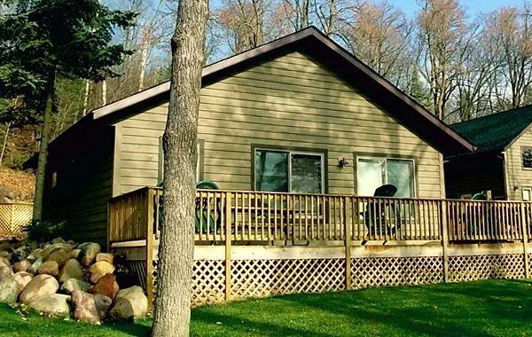 A house with a wooden deck in a backyard, surrounded by trees, with rocks on one side and a grassy lawn in the foreground.