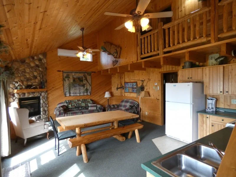 Interior of a log cabin living area with wooden walls and ceiling, featuring a fireplace with stone surround, a dining table with benches, various chairs, a couch, and a refrigerator, with a loft area above and ceiling fans.