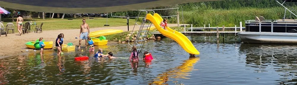 People, including children, swimming and playing near a small beach with a water slide on the shore of a lake. There are paddle boats and boats docked nearby, with chairs and tables under a canopy.