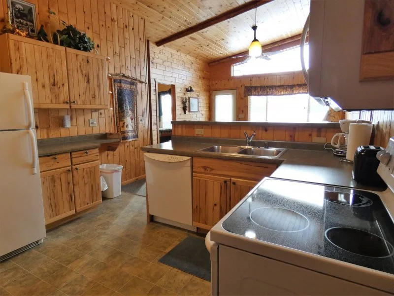 A rustic kitchen with wooden cabinets and walls, white appliances, a double sink, an electric stove, and a small window with a valance. There is a ceiling fan and natural light coming in from windows.