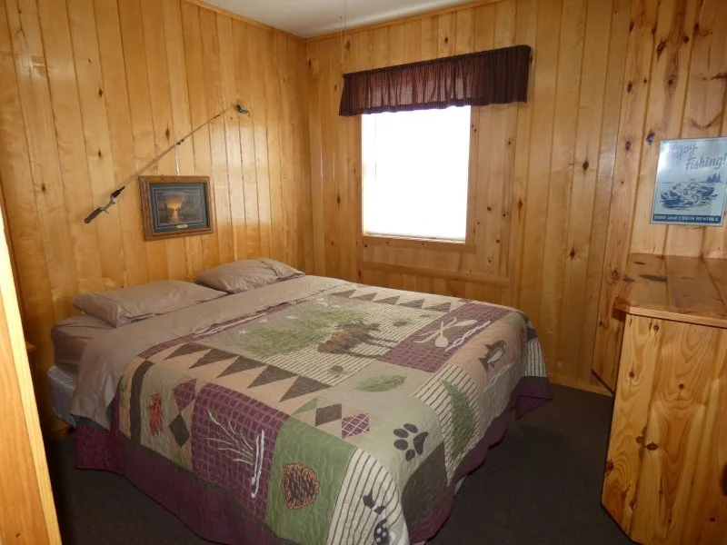 A cozy bedroom with wood-paneled walls and a bed topped with a patchwork quilt and pillows. There is a window with a brown valance, a picture hanging on the wall, and a small wooden dresser to the right.