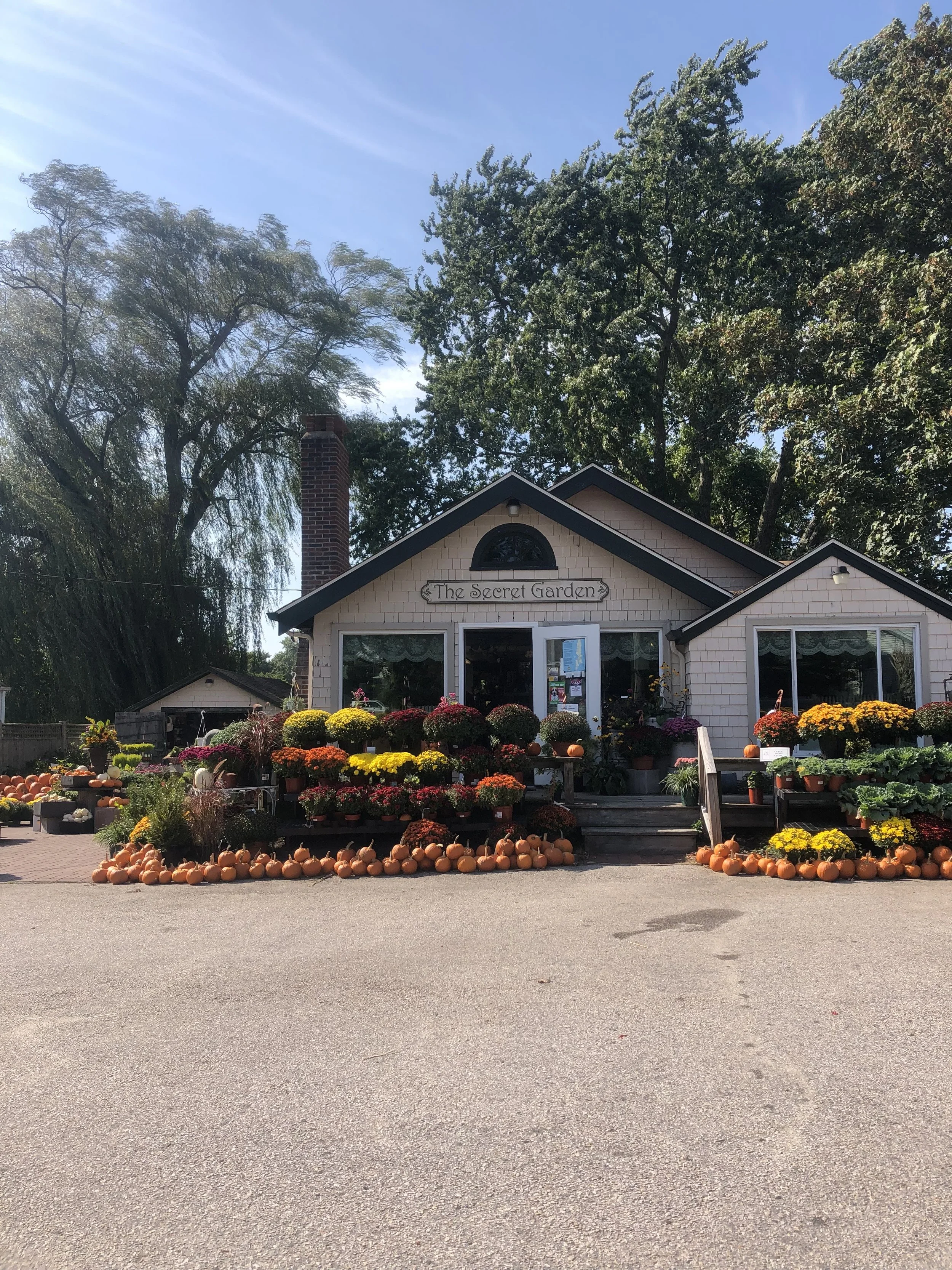 A small building called 'The Secret Garden' decorated with a variety of colorful potted flowers and pumpkins in front, with tall trees and blue sky in the background.