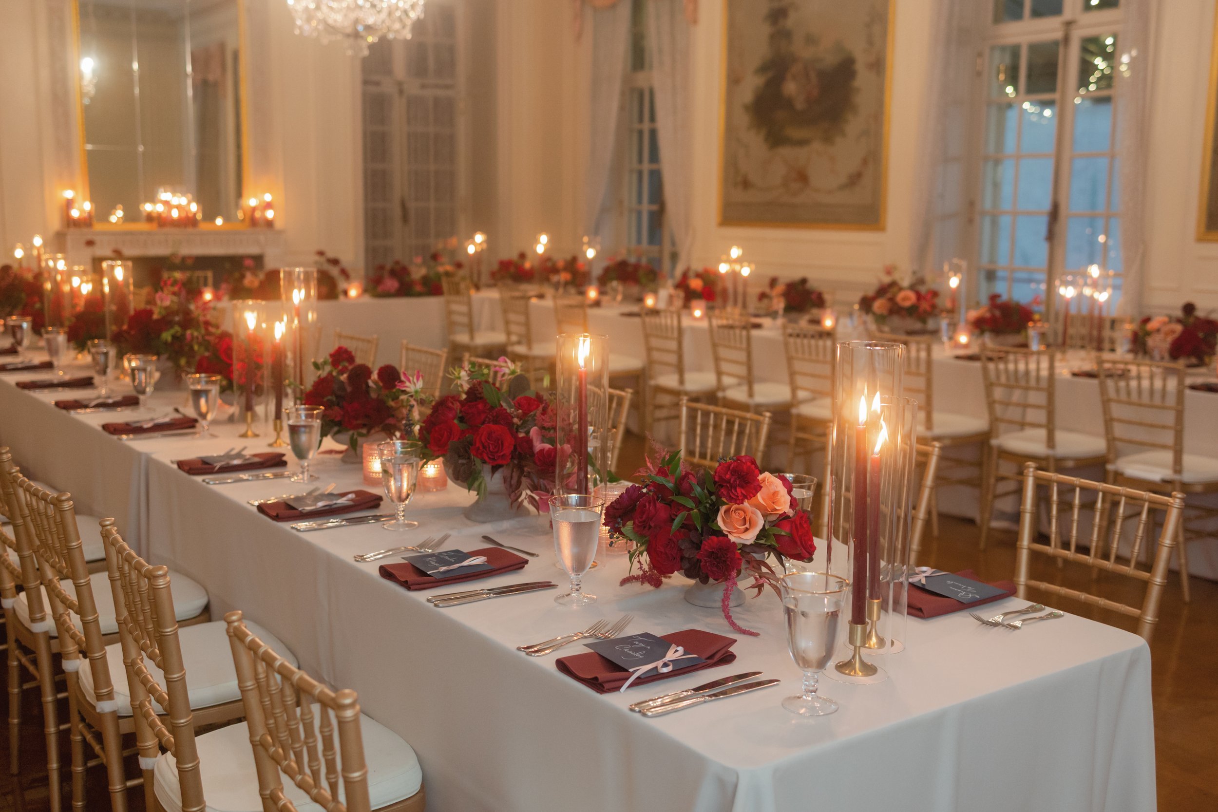 Elegant dining room set up with long table, white tablecloth, floral centerpieces, tall candles, and place settings, in a richly decorated room with large windows and chandeliers.