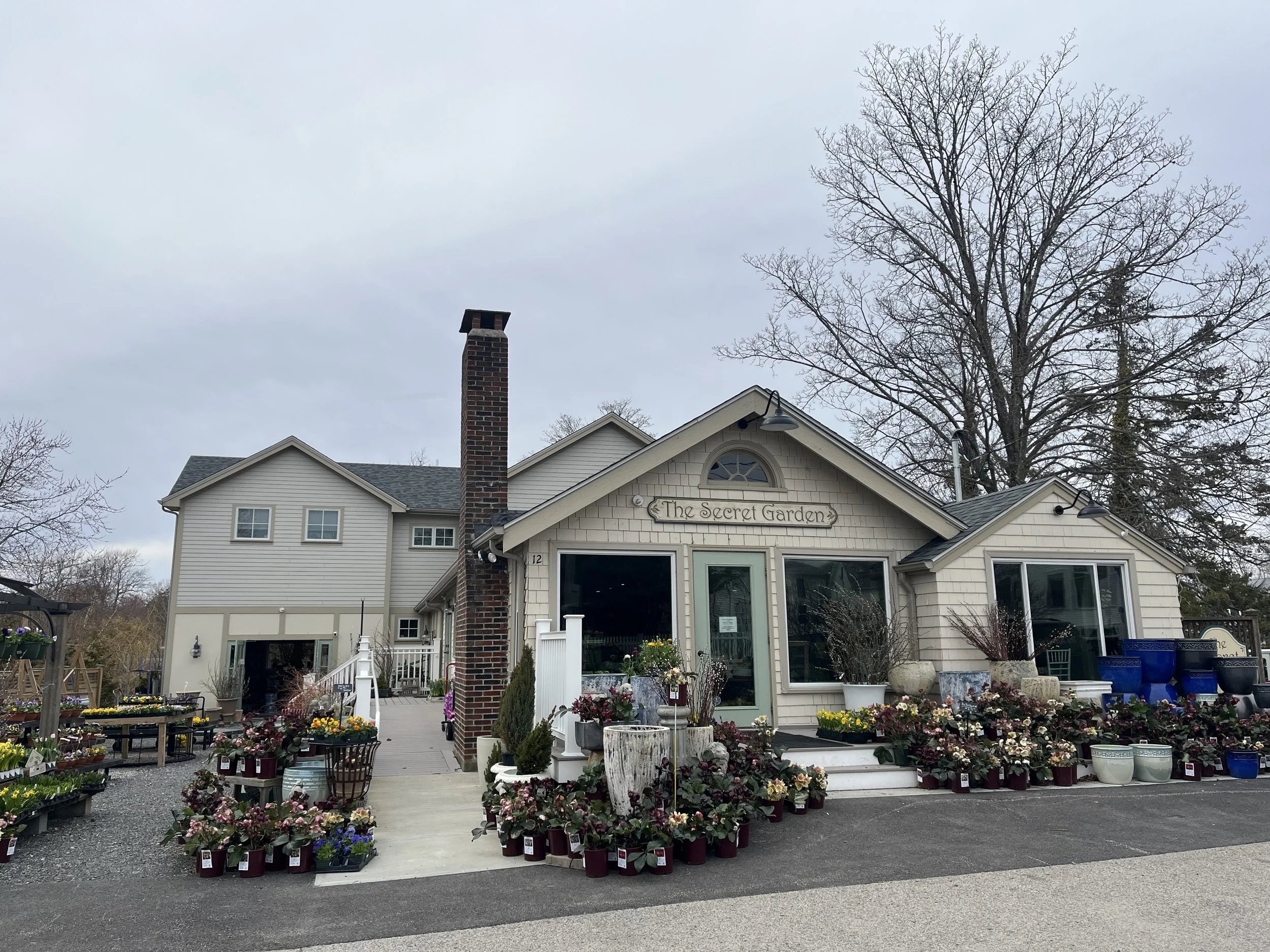 A house with a sign that says "The Secret Garden" above the entrance, with a display of various potted plants and flowers in front of it, and leafless trees in the background.