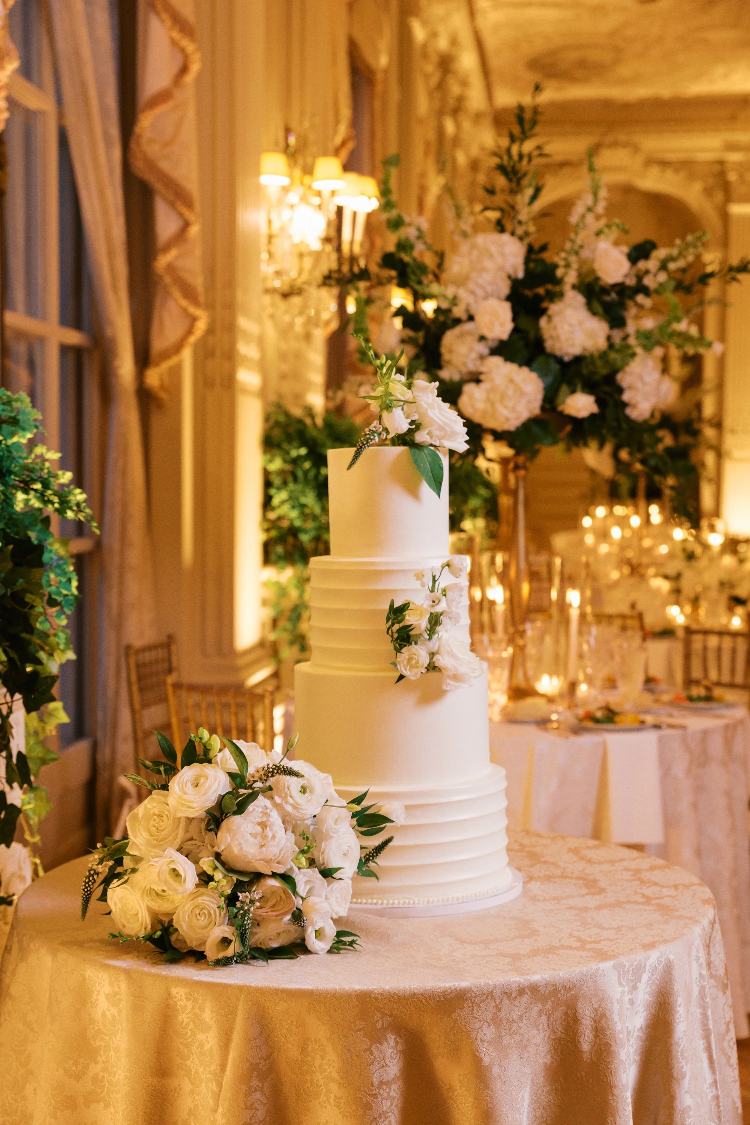 A multi-tiered white wedding cake decorated with white flowers and greenery, placed on a table with a floral arrangement and a beige tablecloth, in an elegantly lit room with draped curtains and floral arrangements in the background.