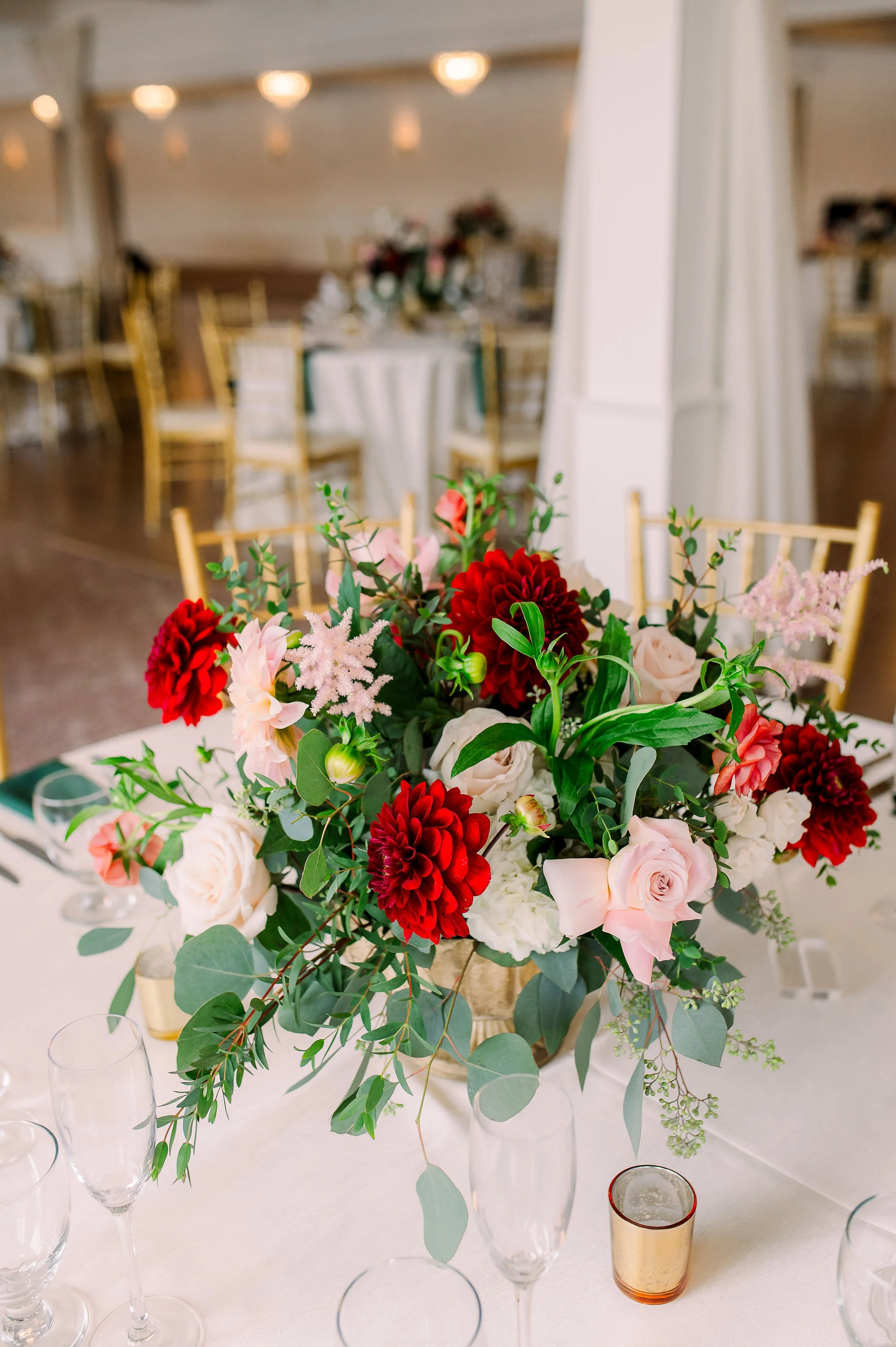 A floral centerpiece with red, pink, and white flowers, surrounded by greenery, on a table with empty champagne flutes and small votive candles.