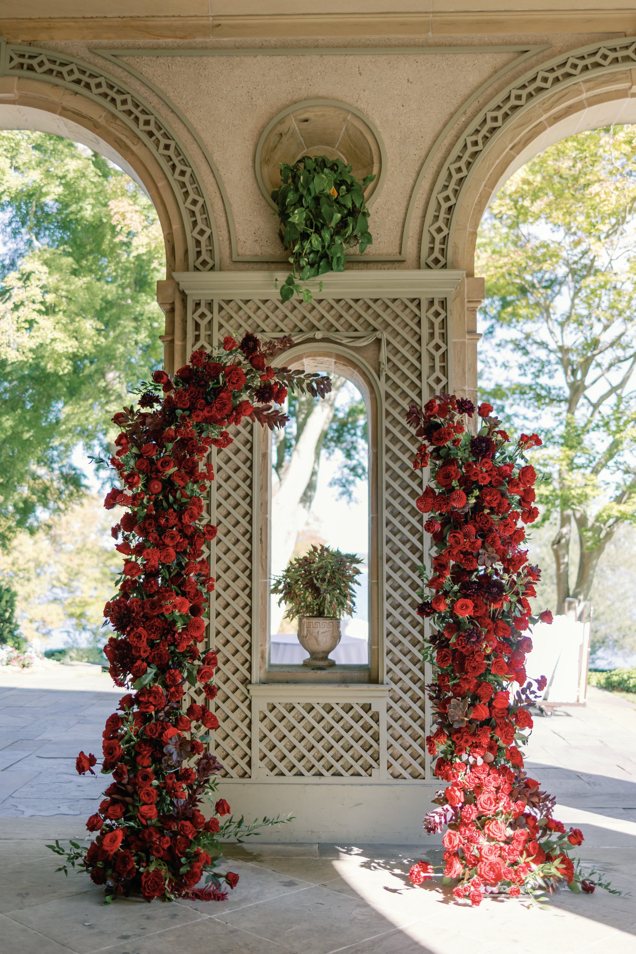 A decorative wedding arch decorated with red and pink roses and dark purple flowers, with a potted plant in a stone vase in the background, under a beige stone pergola with decorative trim, trees and bright sky visible beyond.