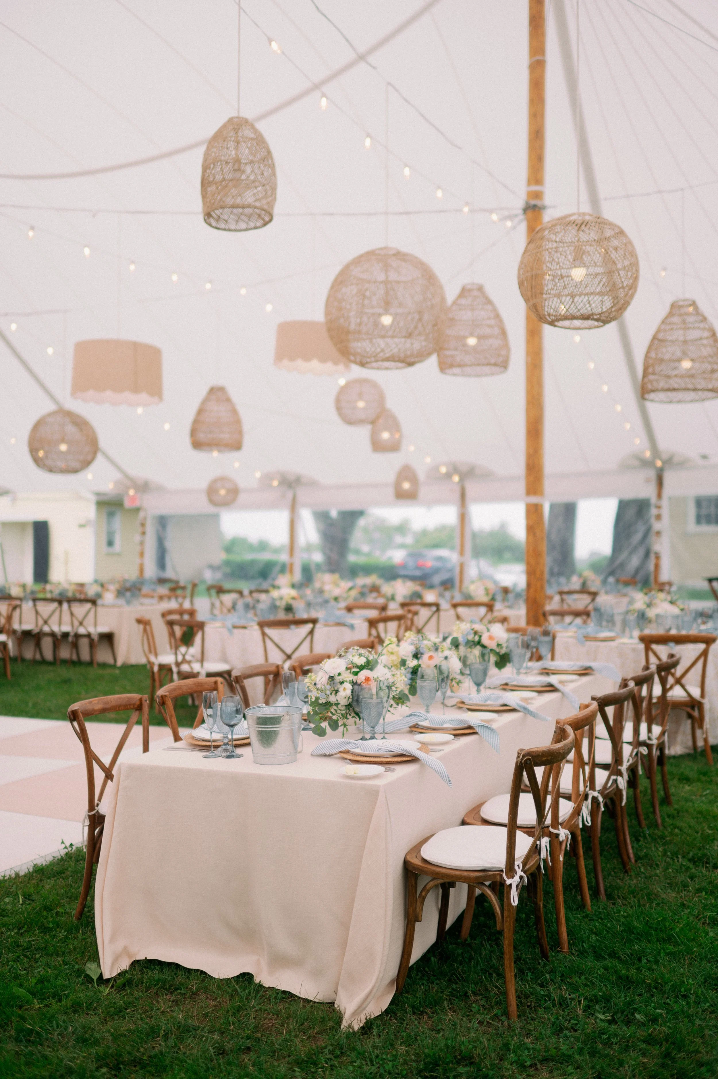 Wedding reception setup under a large tent with tables decorated with white tablecloths, floral centerpieces, and place settings, hanging wicker lanterns, and string lights.