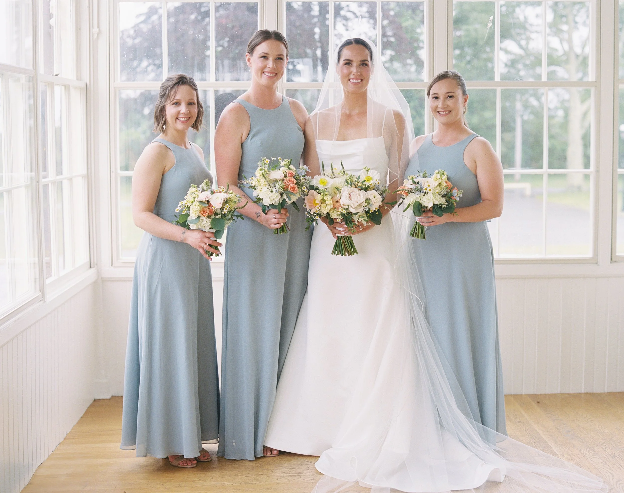 Four women, one bride in a wedding dress with veil, and three bridesmaids in matching light blue dresses, standing together indoors in front of a large window with a view of trees outside. They are holding bouquets of flowers and smiling.