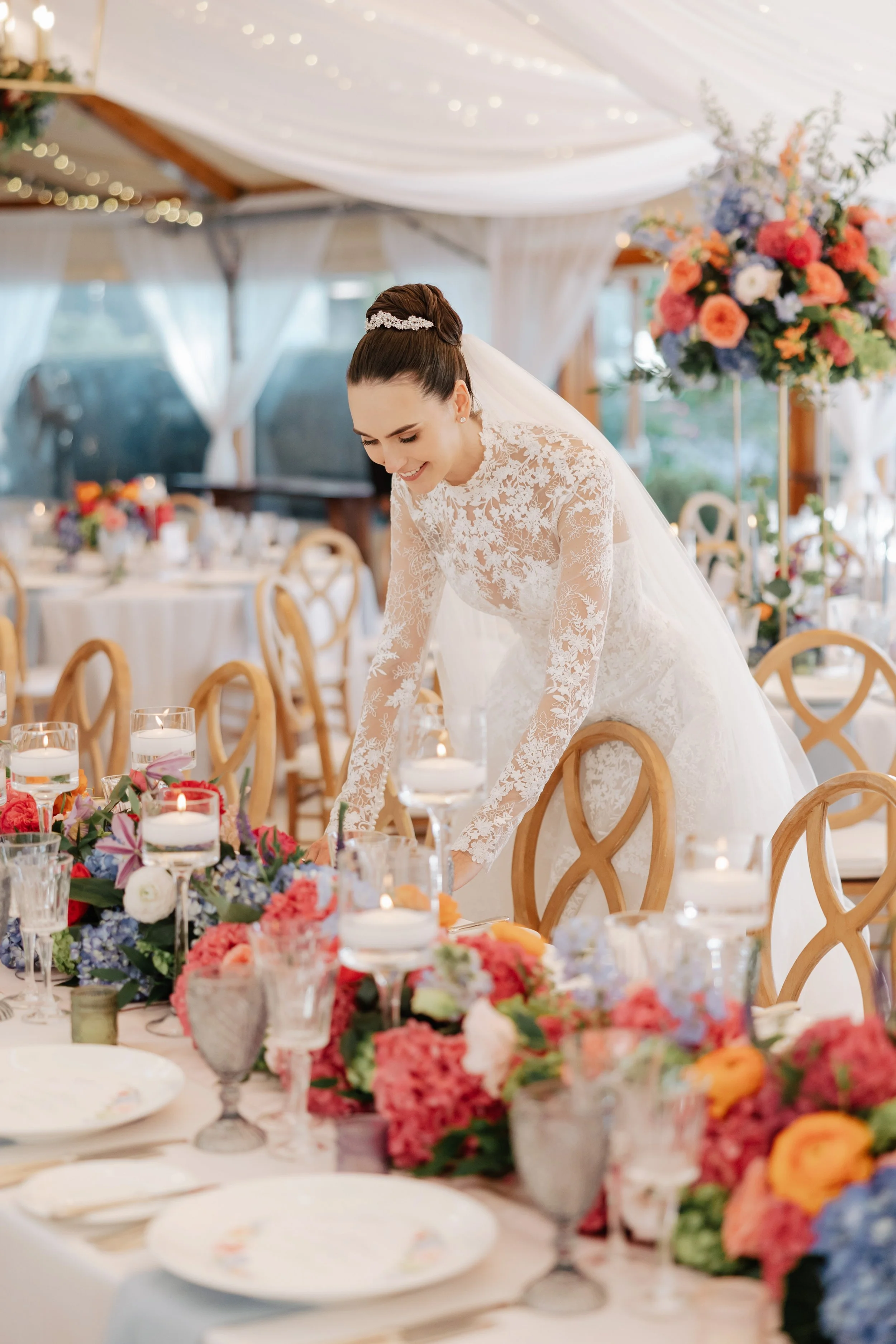 A bride in a lace wedding gown is leaning over a decorated table at her wedding reception. The table is adorned with colorful flowers and candles. The setting appears to be a decorated indoor venue with white drapes and fairy lights.