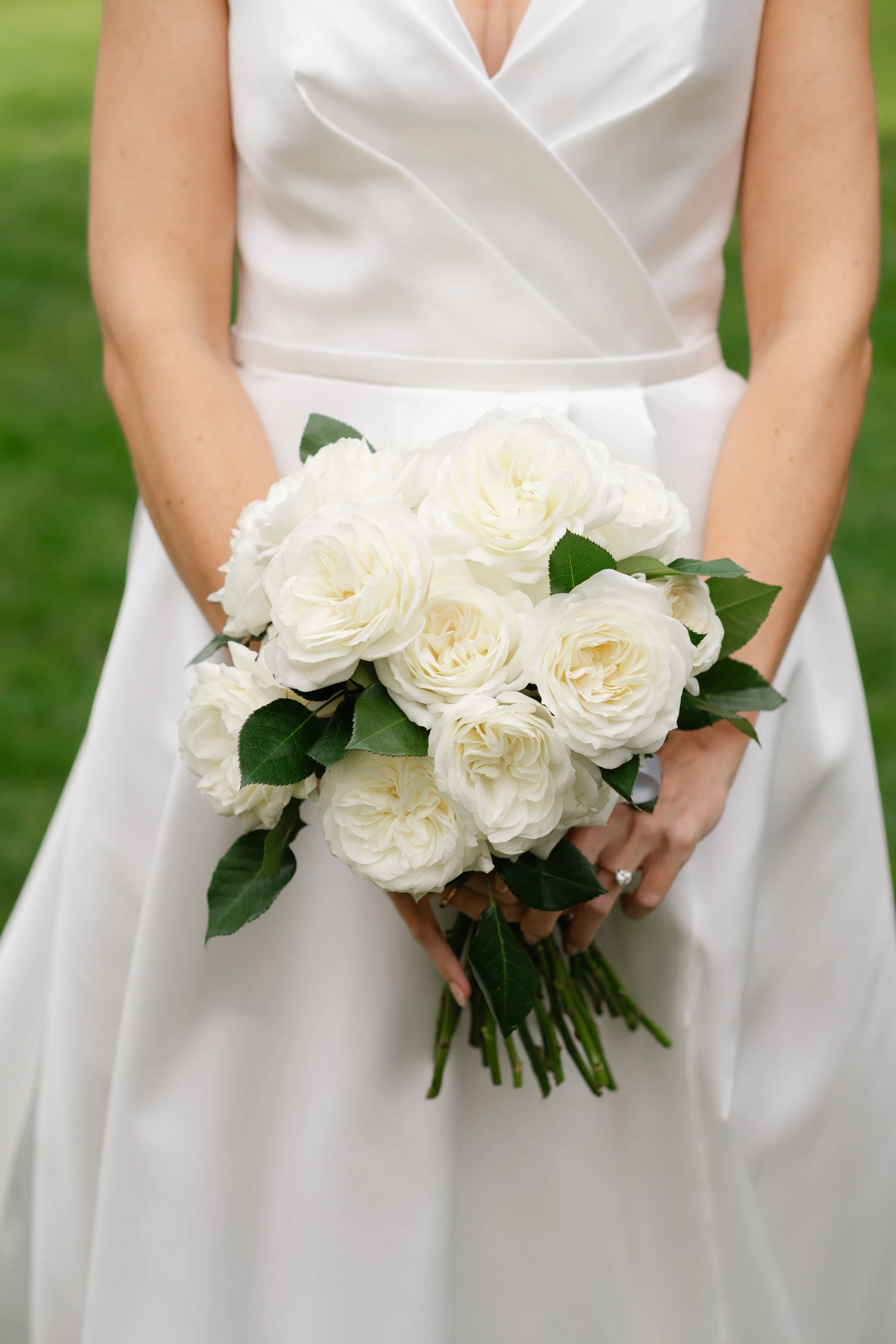 A woman in a white dress holding a bouquet of white roses with green leaves, standing outdoors with a blurred green background.