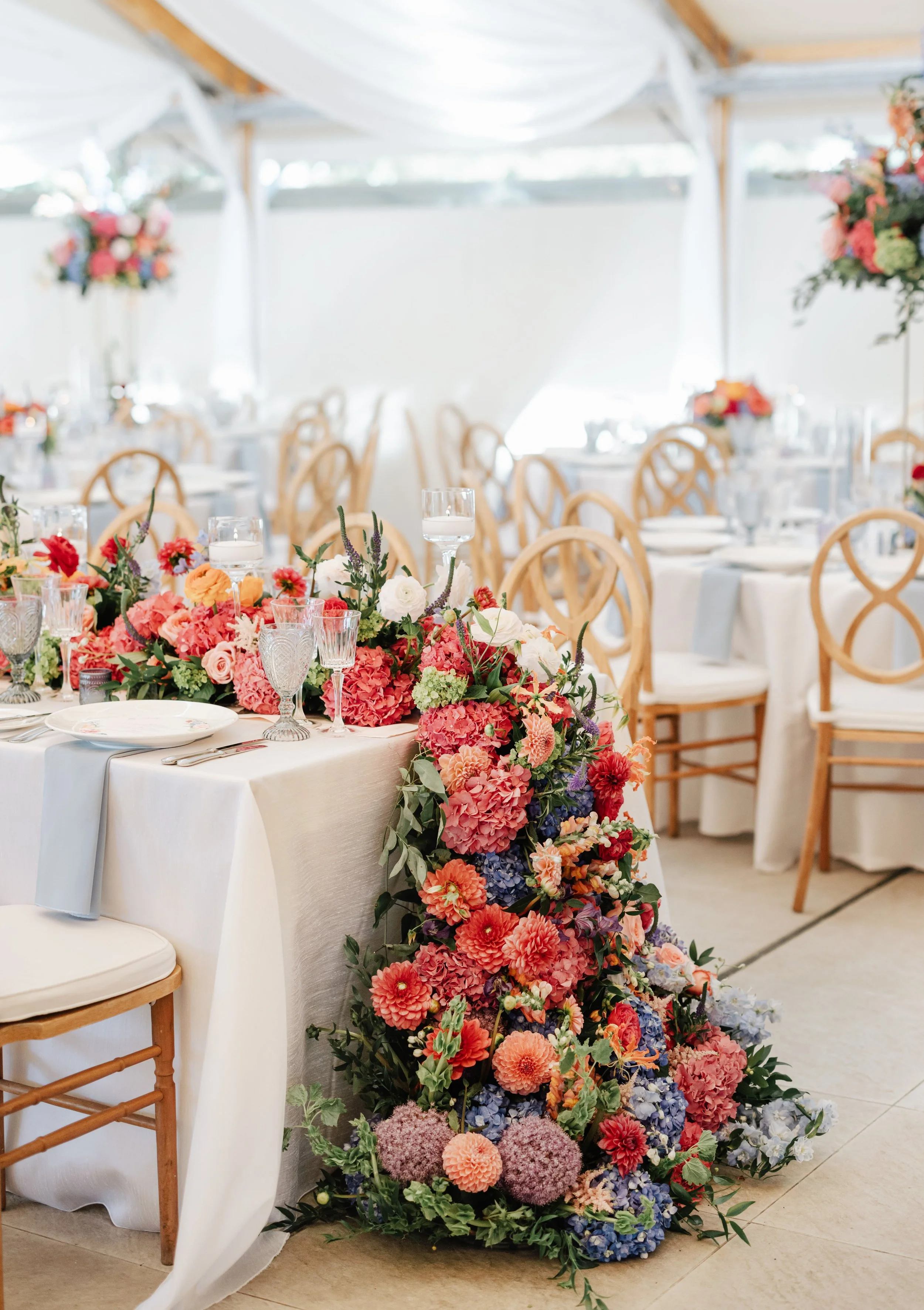 A wedding or event table decorated with a large cascading floral arrangement featuring pink, red, purple, blue, and white flowers. White tablecloth, glassware, plates, and silverware are set on the table, and wooden chairs surround it in a bright, white tent with draped fabric.