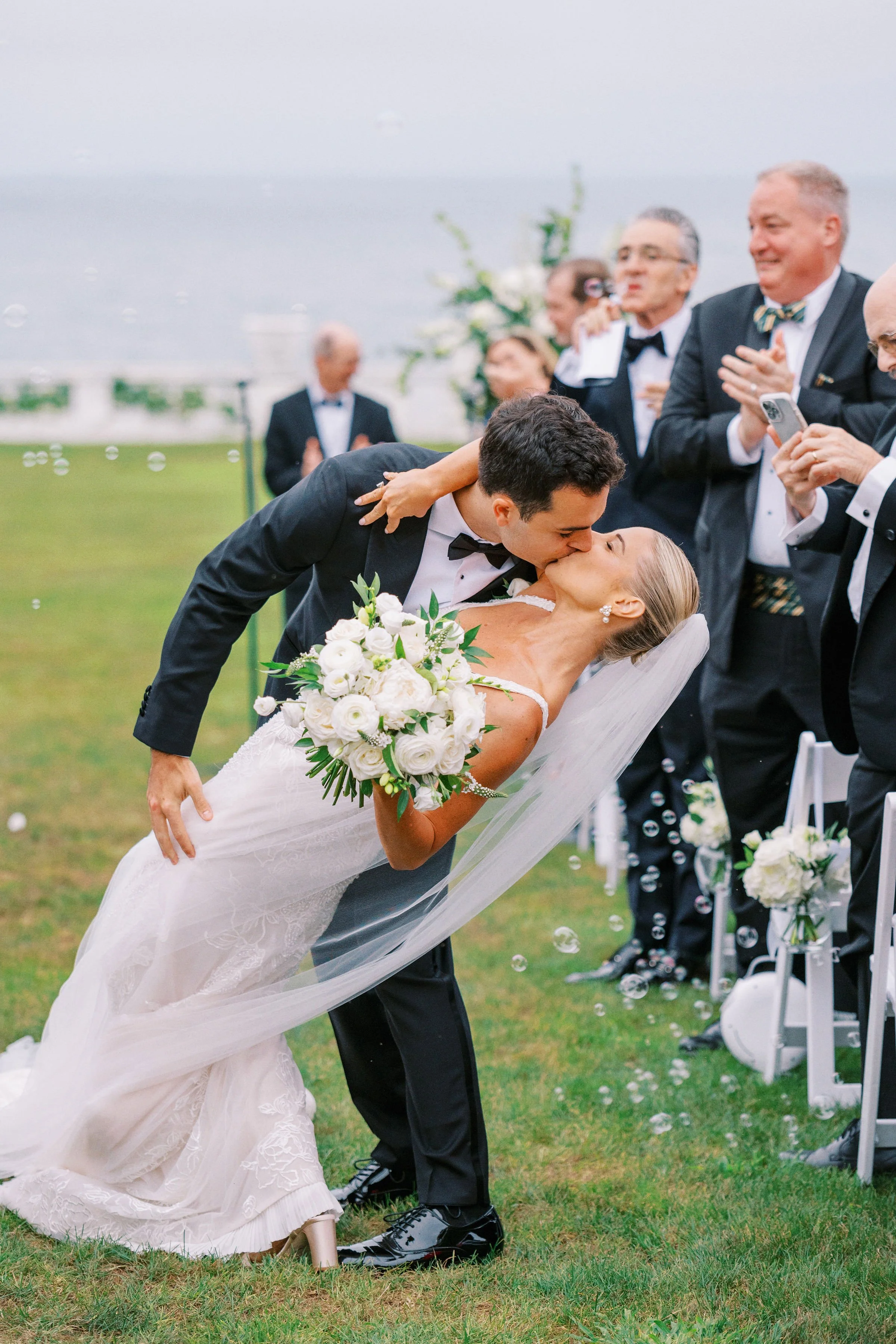 A bride and groom, dressed in wedding attire, share a kiss during their outdoor wedding ceremony on a grassy field near the ocean, with guests clapping and guests using phones to capture the moment.