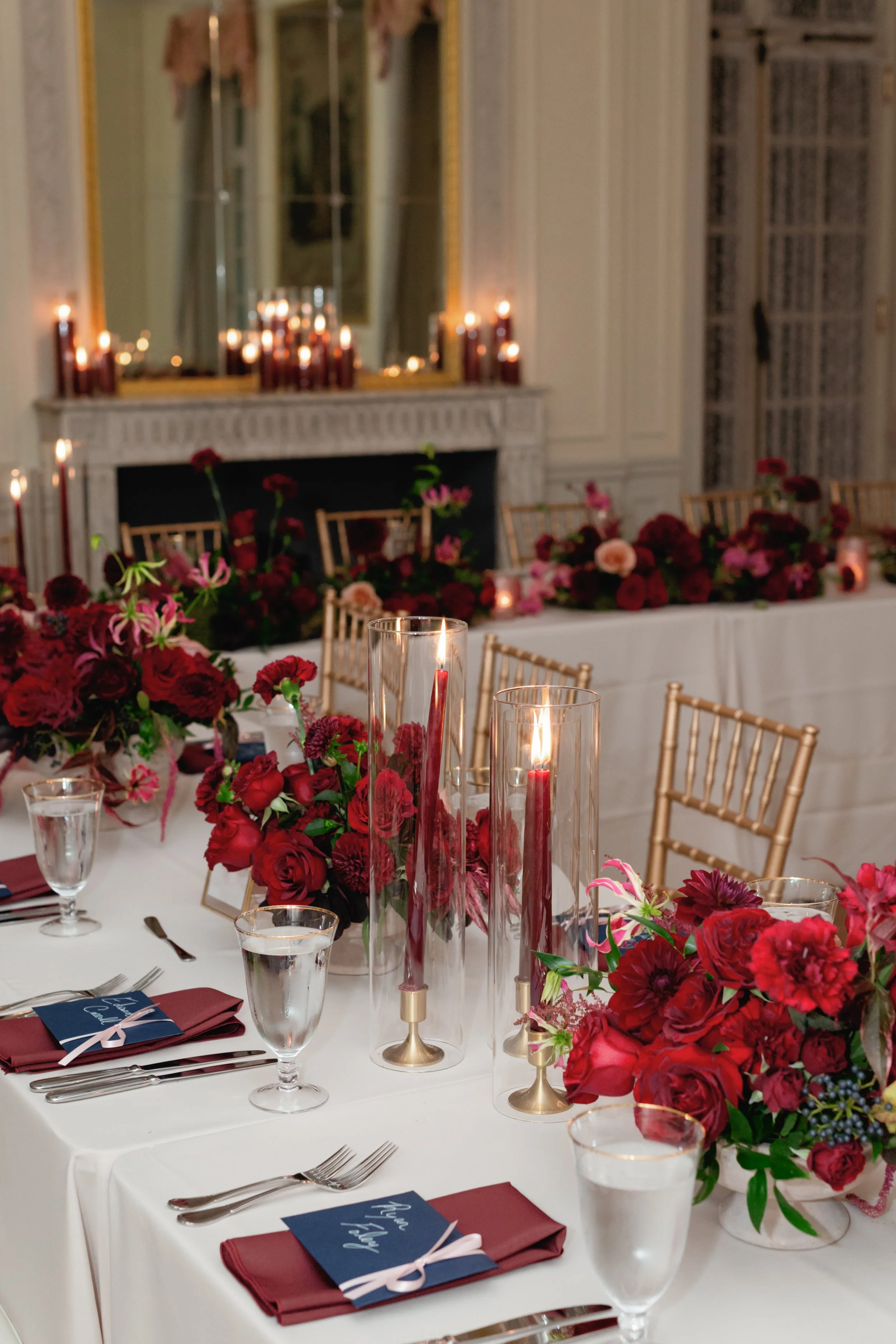 Elegant dining table with floral centerpieces, candles, and place settings in a decorated room with a fireplace and mirror.