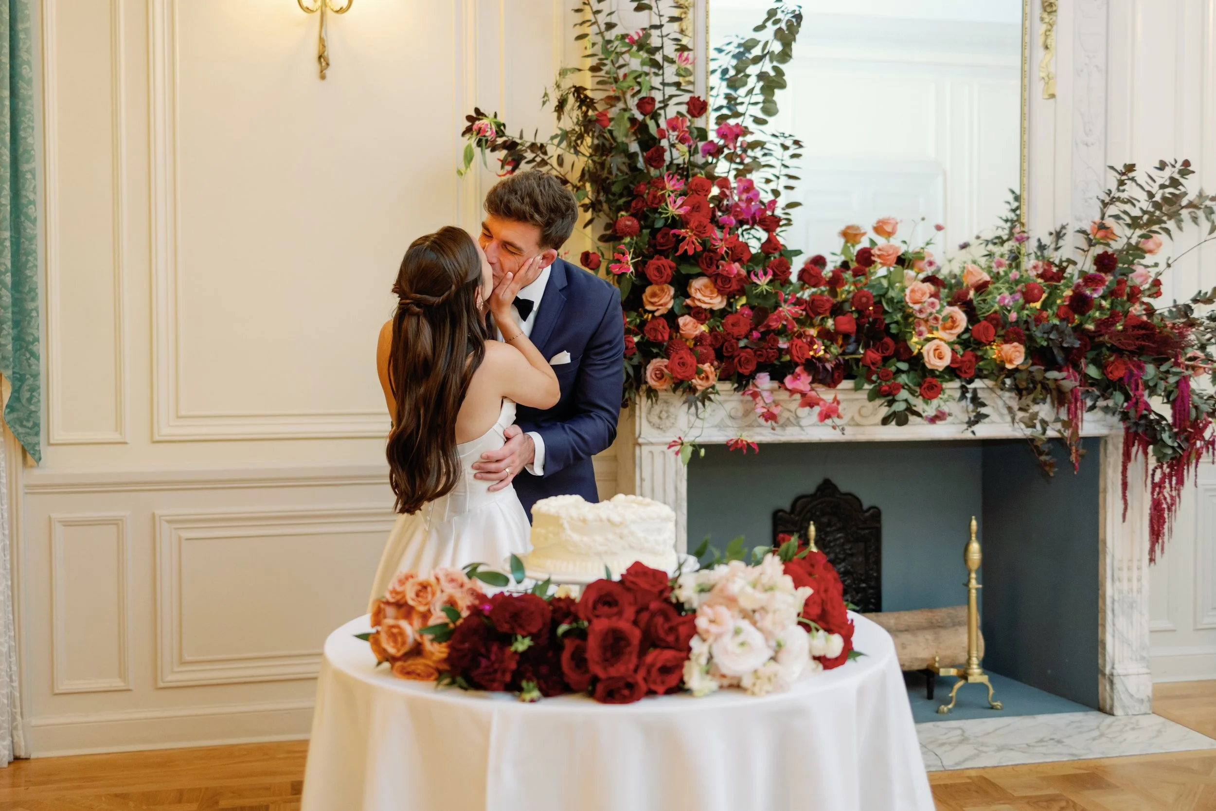 A couple kissing at a wedding reception, with a decorated table and floral arrangement in the background.