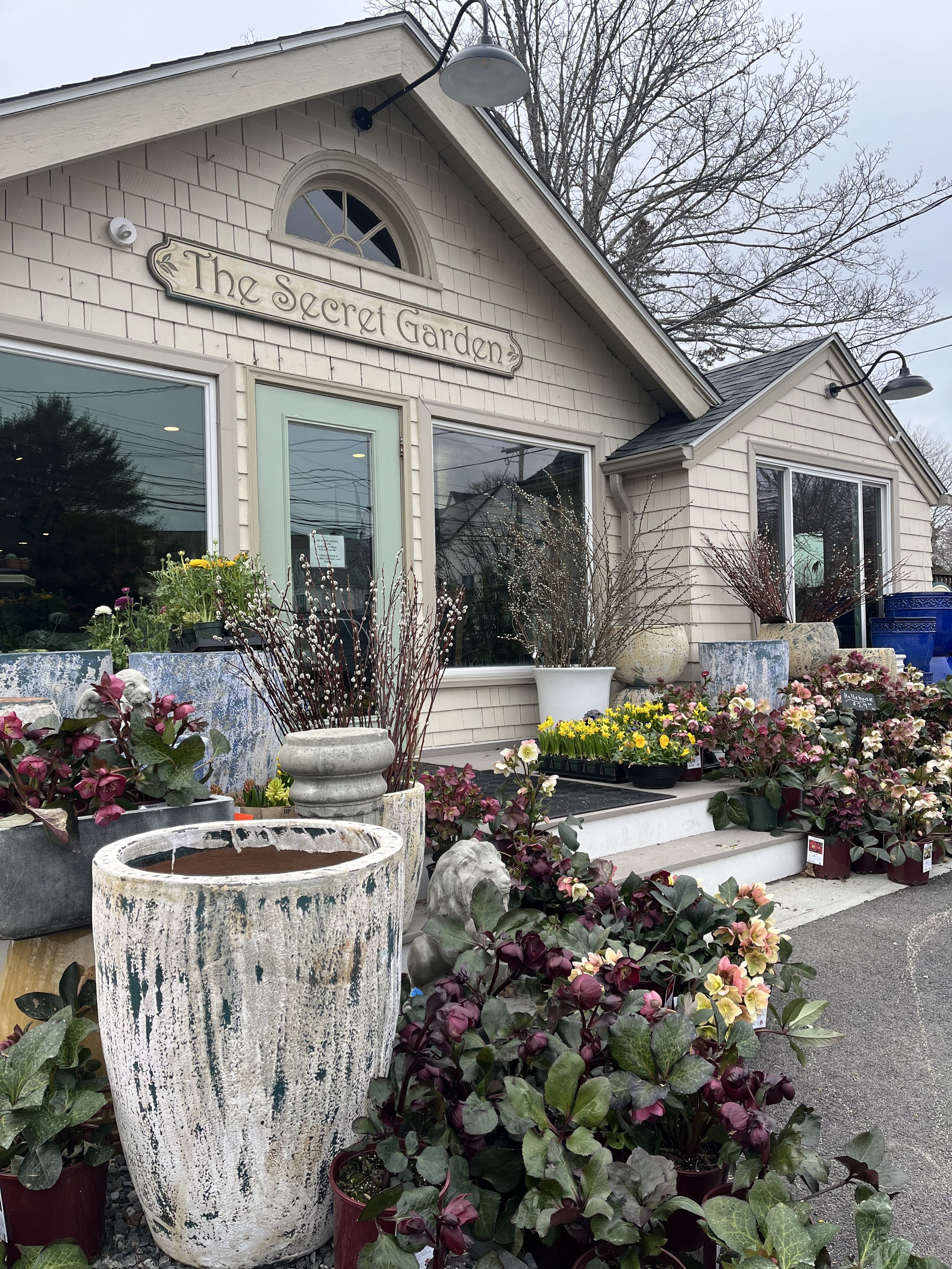 An outdoor scene at a garden shop titled 'The Secret Garden'. The shop has large windows, and the foreground is filled with various potted plants, flowers, and decorative garden items, including large ceramic pots and a wooden sign.