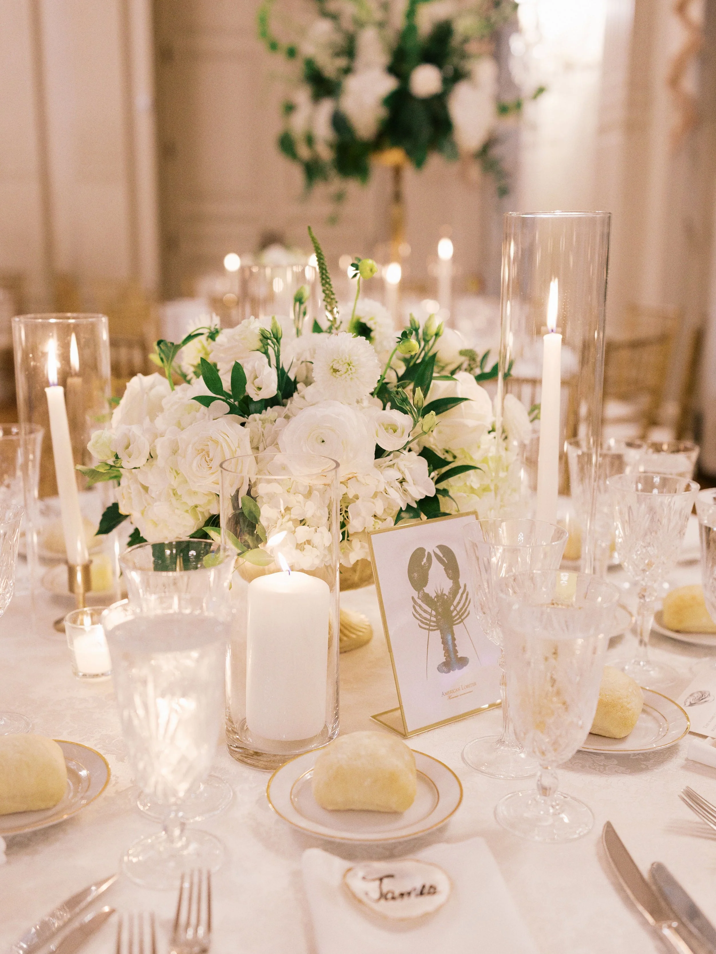 Elegant dining table decorated with white flowers in a clear vase, surrounded by glass candle holders with lit candles, and a place card labeled 'James'.
