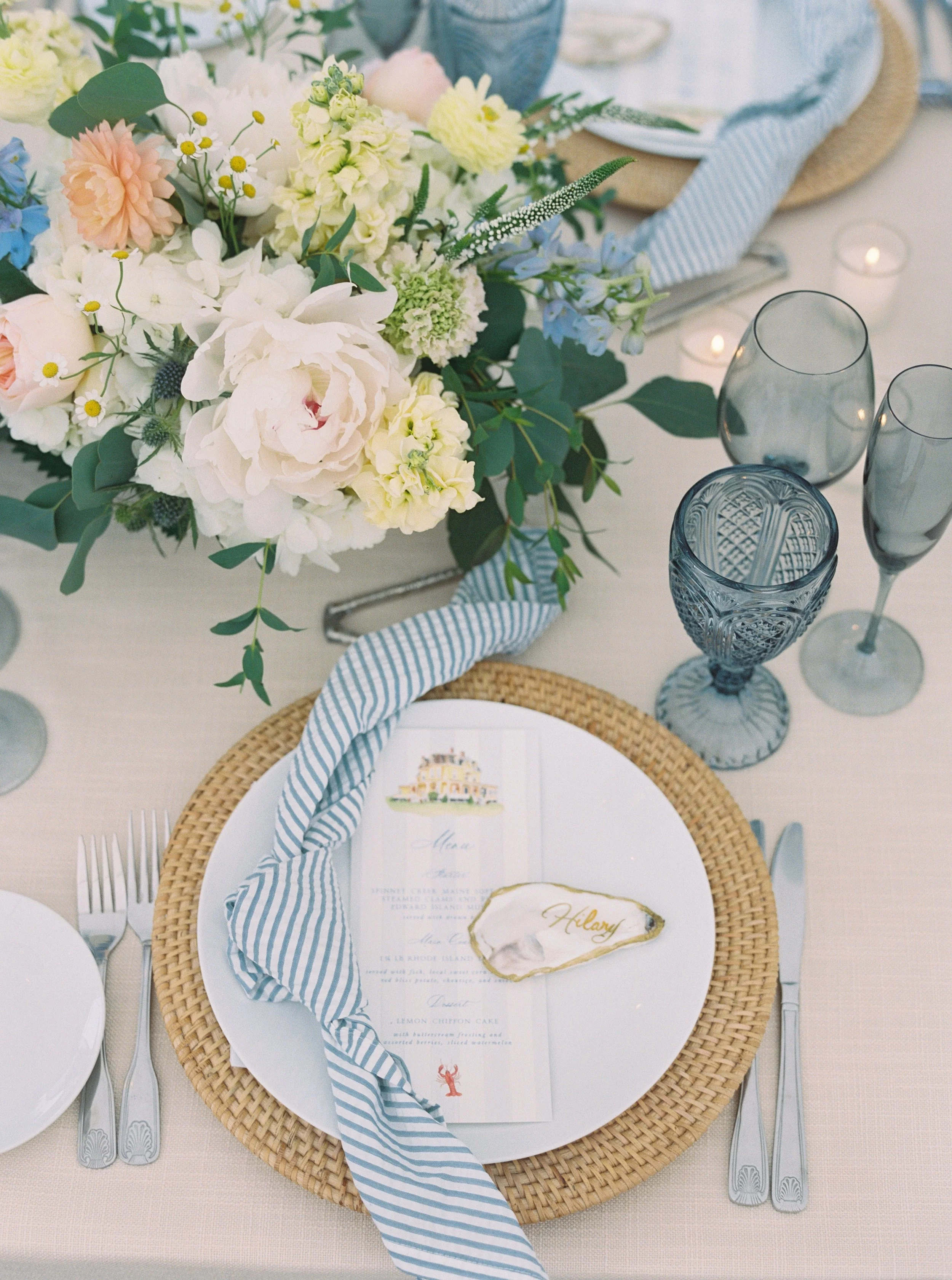 A fancy table setting with a floral centerpiece, a menu card, and place setting. The centerpiece contains white, cream, and pink flowers with greenery. The place setting includes a white plate on a woven placemat, a blue and white striped napkin, and silver cutlery. There are blue glasses and a water glass. The menu card has a gold-rimmed shell with the name 'Hilary.' Small candles are on the table in the background.