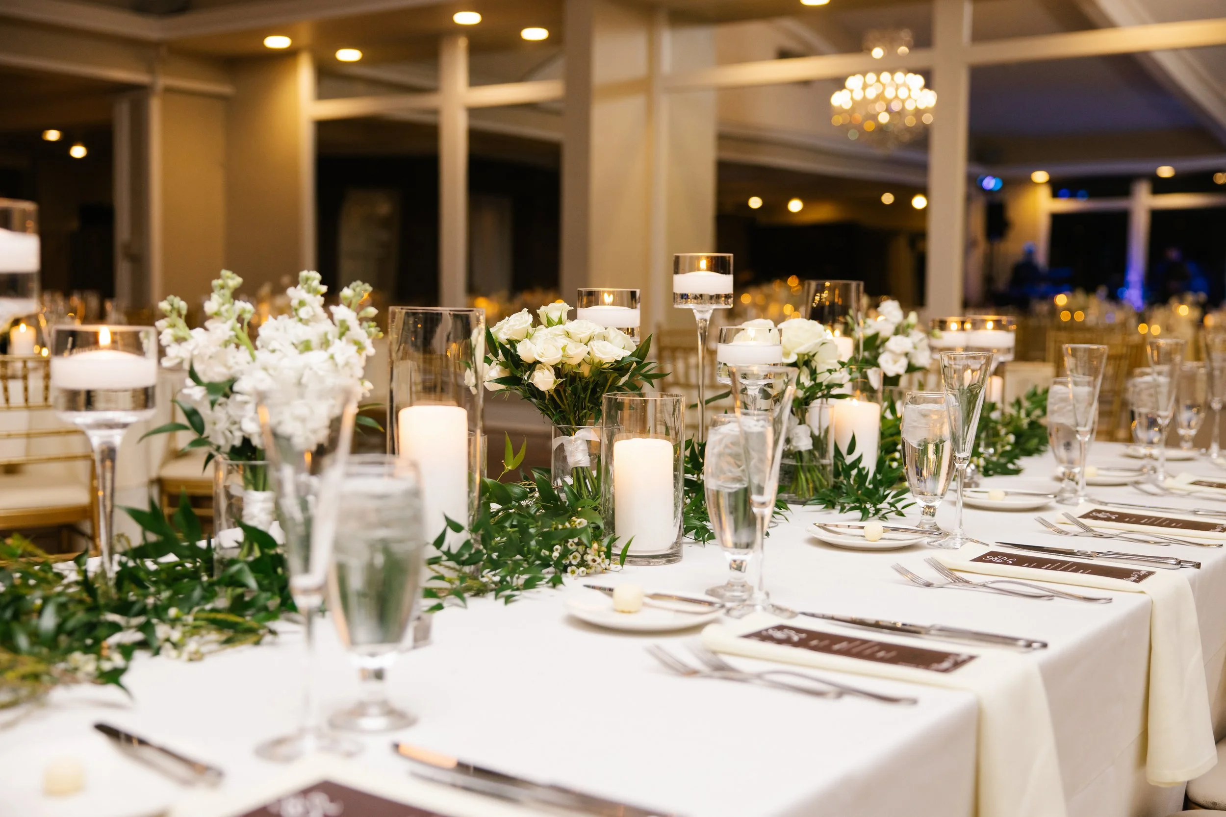 Elegant banquet table decorated with white flowers, candles, and glassware in a well-lit reception hall.