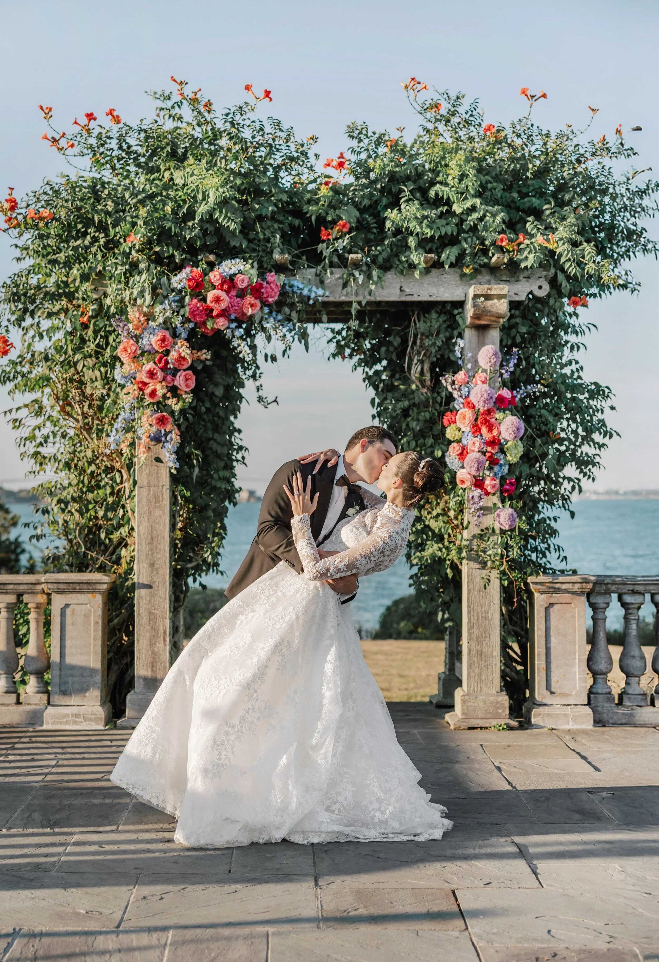 A bride and groom sharing a kiss under a floral arch by the water during their wedding ceremony.
