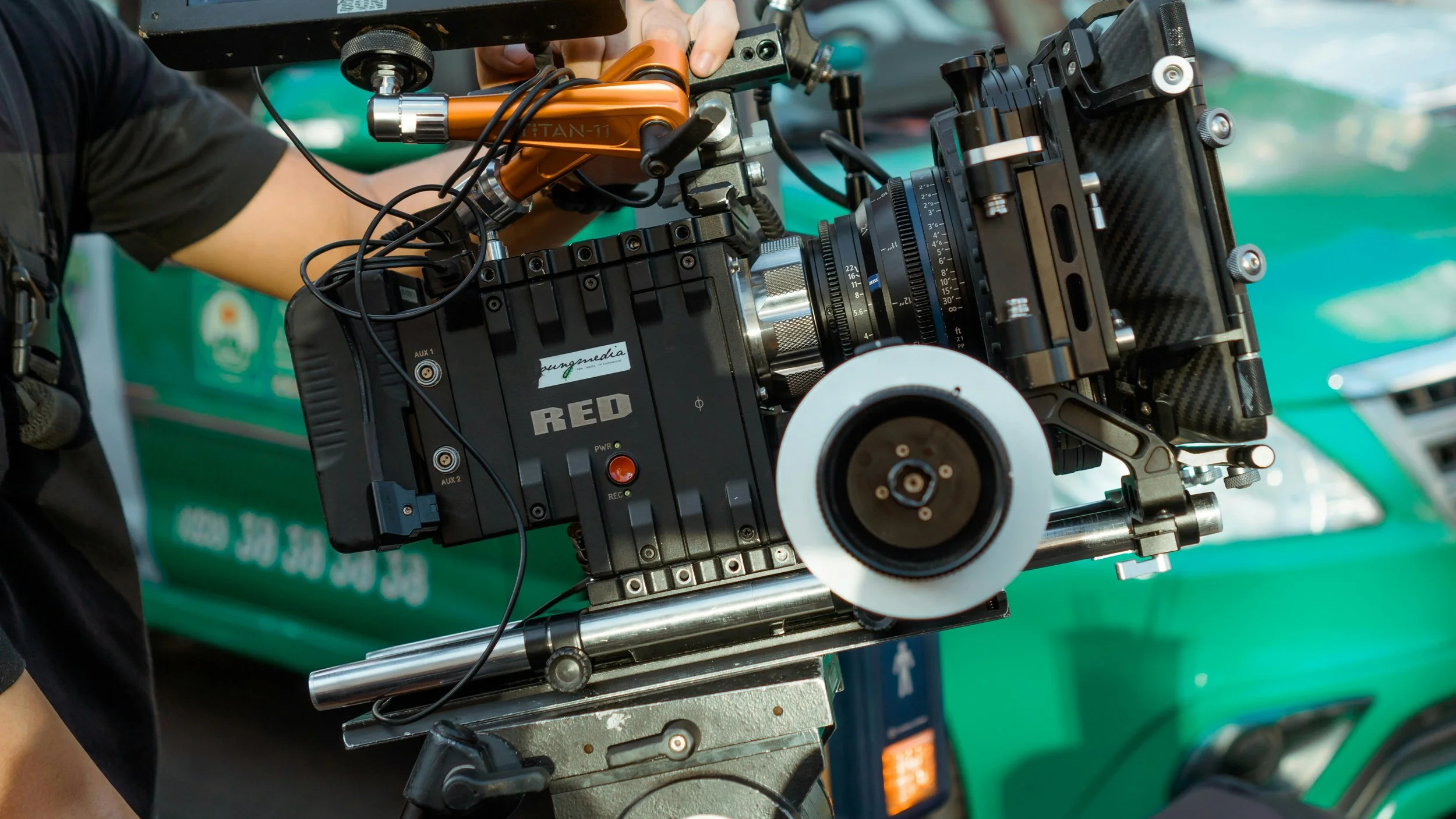 Close-up of a professional film camera on a dolly, with a person adjusting it, showing various controls, cables, and camera components.