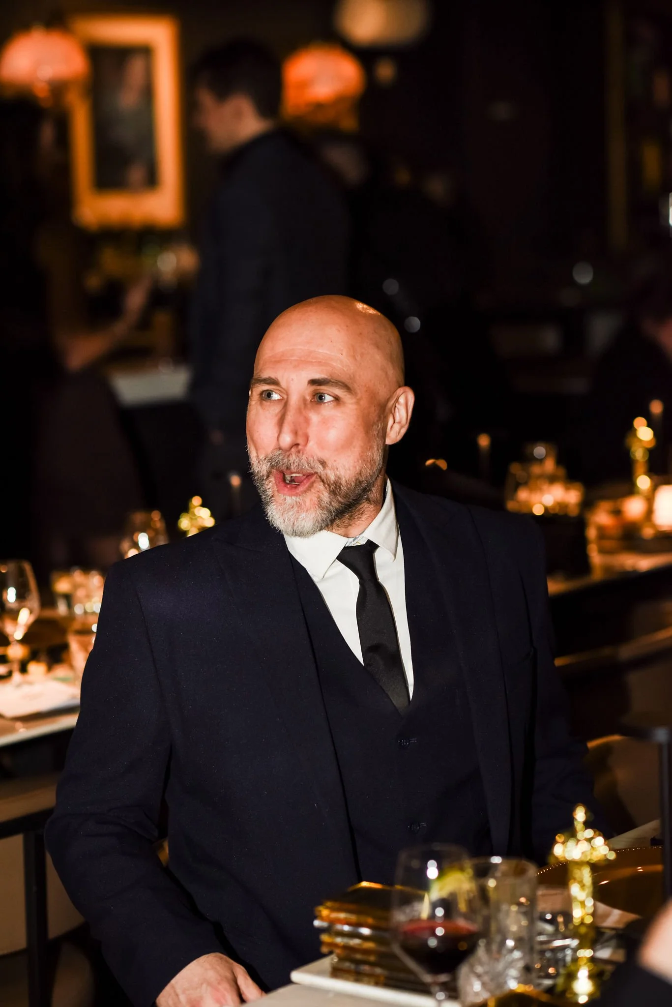 A man with a bald head, gray beard, and light skin, wearing a black suit and tie, is sitting at a restaurant or bar. The background shows other people, including a man in a black outfit, and warm orange lighting, indicating a cozy, upscale setting.