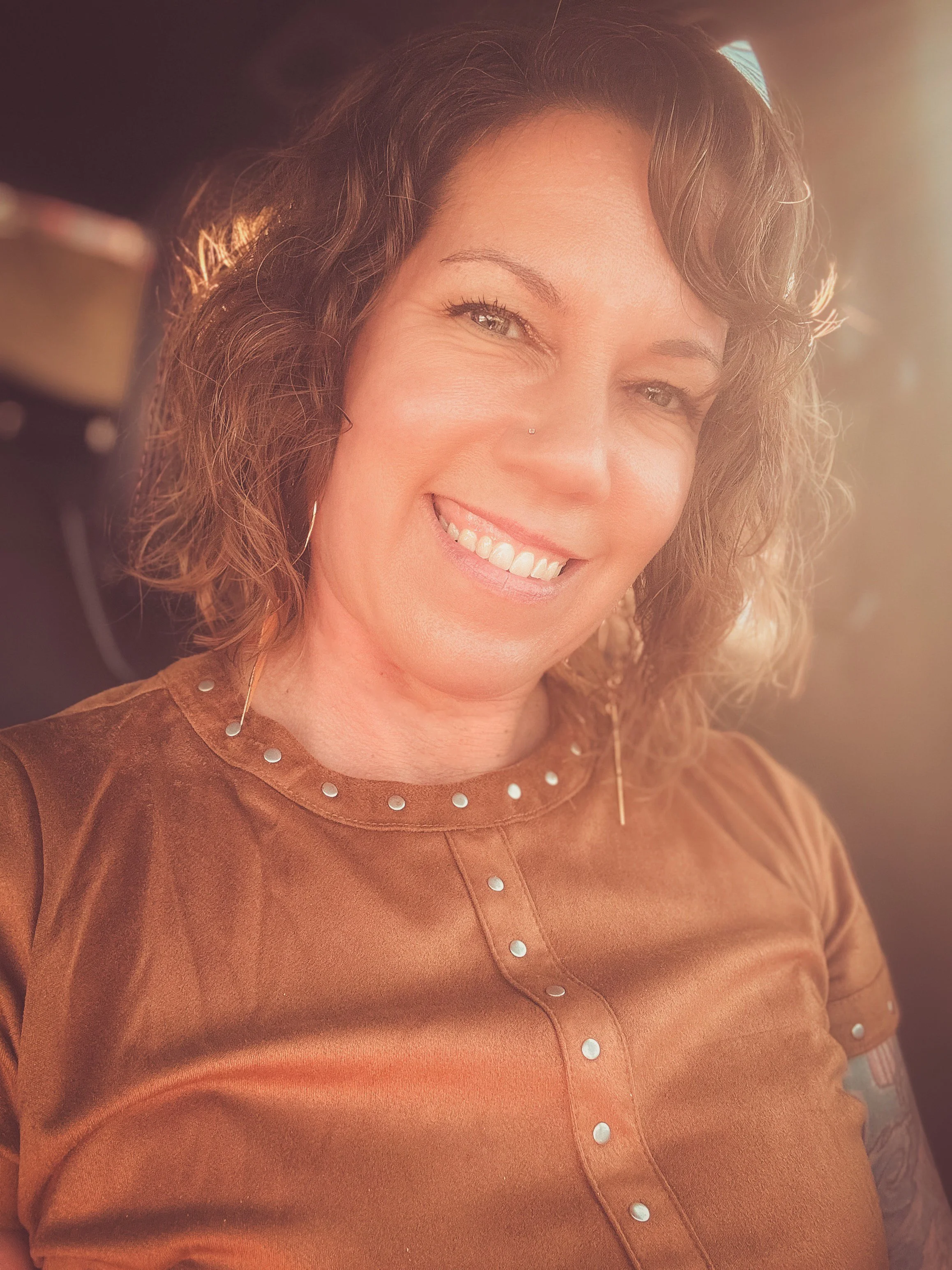 A woman with curly brown hair smiling at the camera, wearing a brown top with silver embellishments around the collar and down the front, and a nose ring.